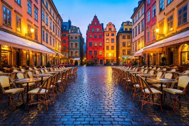 Op een kasseiplein staan bij het vallen van de avond cafétafels en stoelen tussen historische gevels in rood, oranje en geel, met warme verlichting onder een diepblauwe hemel.