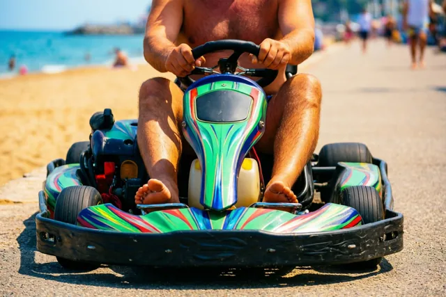 Een man met ontbloot bovenlijf rijdt op een blauwe, groene en gele gocart over een verharde zeedijk, met strand en zee in de zon op de achtergrond.