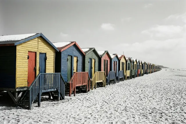 Een rij kleurrijke houten strandcabines met sneeuw op de daken staat op een leeg zandstrand onder een bewolkte hemel.