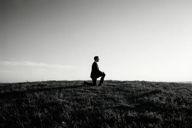Op een zwart-witfoto knielt een man in een pak op één knie op een grasheuvel, als silhouet tegen een heldere hemel.