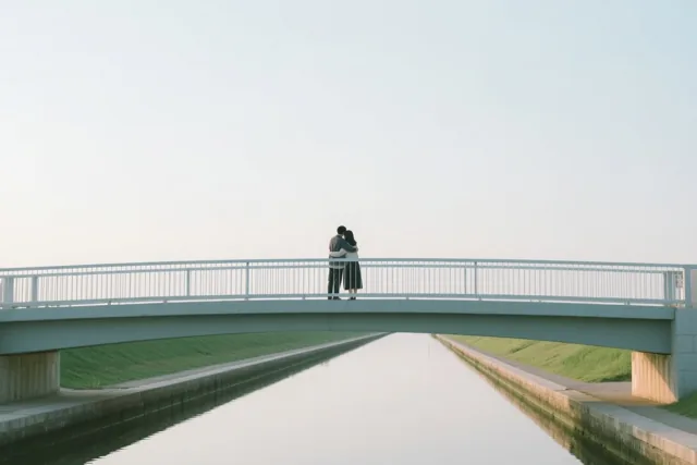 Twee mensen omhelzen elkaar op een witte voetbrug boven een rustig kanaal met groene grasoevers onder een bleke hemel.