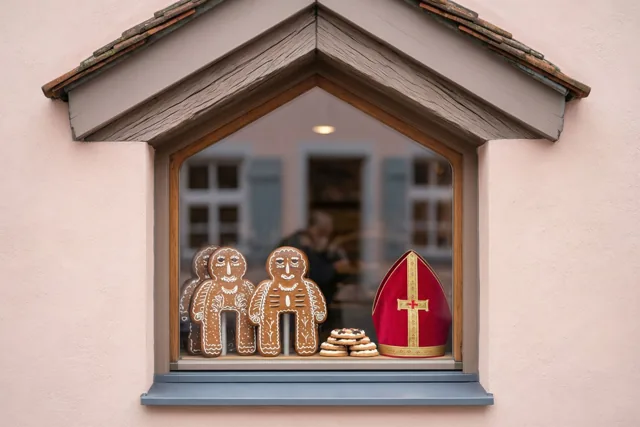 Een warm verlichte bakkersetalage onder een trapgevel toont peperkoeken figuren, een rode mijter en een stapel koekjes tegen een roze muur.