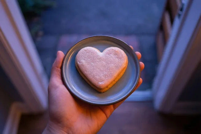 Een hand houdt bij een deuropening een bordje met een hartvormig koekje met glazuur vast, in warm licht tegen een koele achtergrond.