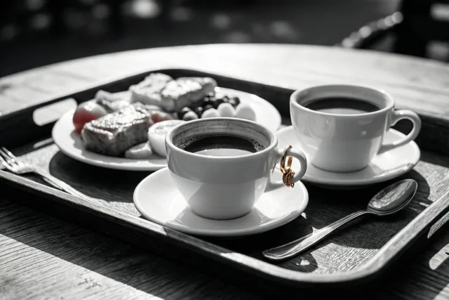 Een zwart-witfoto toont twee koffiekopjes op een dienblad met een ring aan het oor van één kopje, naast een bord gebak en bestek op een zonnige houten tafel.