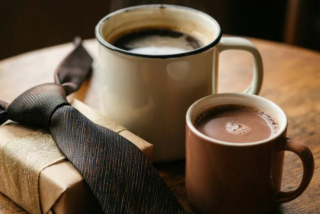 Een grote witte mok koffie en een kleine bruine mok warme chocolademelk staan op een houten tafel naast een pakje in bruin papier, dichtgebonden met een das met patroon.
