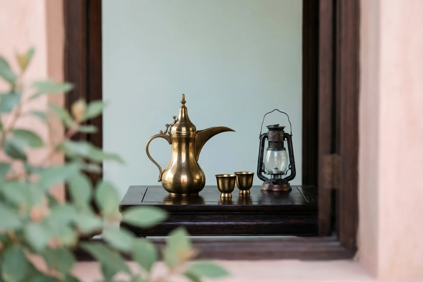 Door een houten raamkozijn zijn een messing dallah-koffiepot, twee kleine kopjes en een maansikkellantaarn op donker hout te zien, met een lichtblauwe muur erachter en groen gebladerte op de voorgrond.