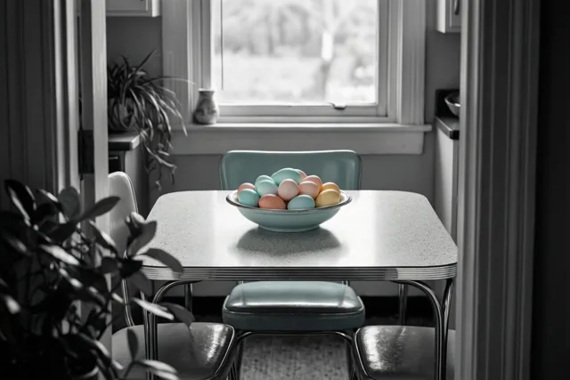 Een kom met pastelkleurige paaseieren staat op een formica keukentafel in jaren-vijftigstijl, in een vintage keuken met gedempte kleuren en daglicht.