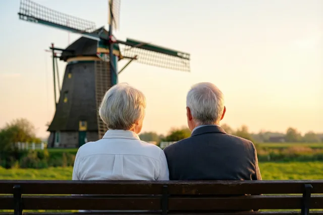 Een ouder koppel zit, van achteren gezien, op een houten bank en kijkt naar een traditionele windmolen in een landelijk landschap bij zonsondergang.