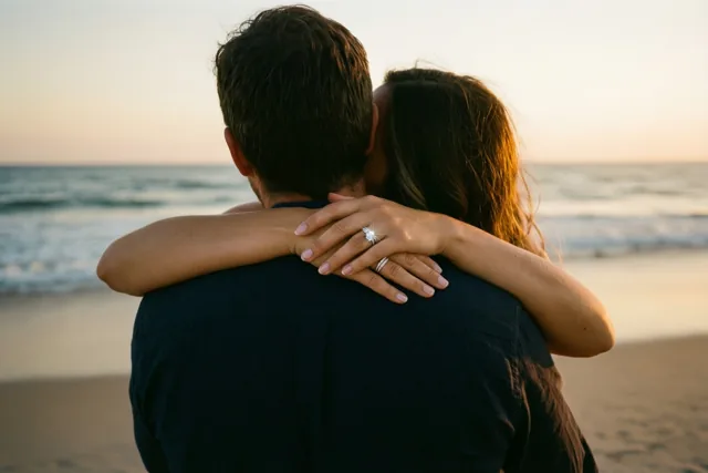 Een koppel omhelst elkaar met de rug naar de camera op een strand in warm gouden schemerlicht, terwijl de hand van de vrouw met een fonkelende ring op de schouder van de man rust en de zee achter hen ligt.