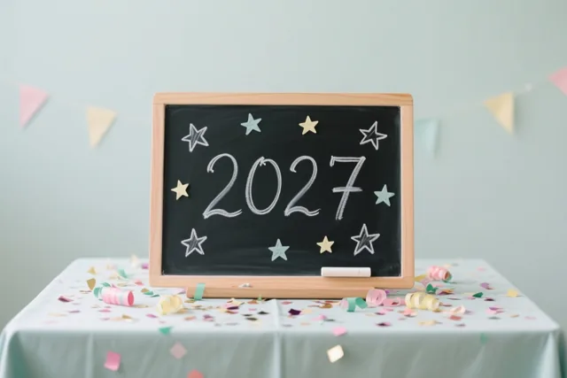 Een klein krijtbord met 2027 en sterren in wit krijt staat op een tafel vol confetti en feestversiering, met een vlaggenlijn voor een lichte muur.