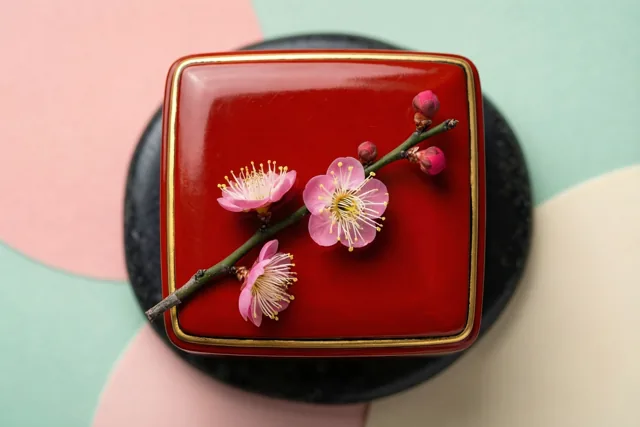 Een rood lakdoosje met gouden rand bevat een takje roze pruimenbloesem en rust op een zwarte steen tegen een roze, mintgroene en crèmekleurige achtergrond.
