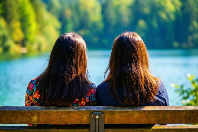 Twee mensen met lang haar zitten met hun rug naar de camera op een houten bank aan een rustig meer, met groene bomen in fel zonlicht op de achtergrond.