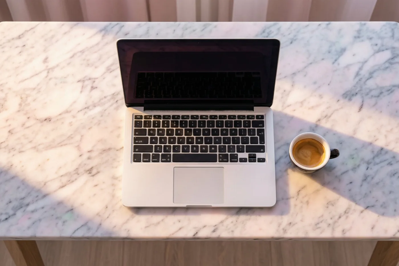 Een zilverkleurige laptop en een wit espressokopje staan op een wit marmeren bureau in warm ochtendlicht.