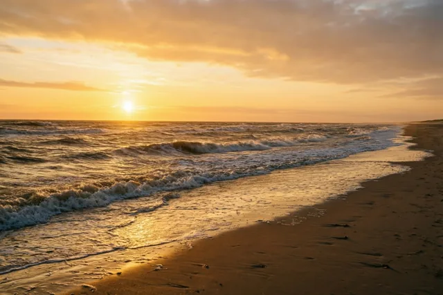 Zachte golven rollen op een breed, leeg strand bij zonsondergang, met gouden en oranje tinten boven de zee.