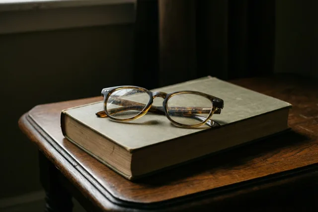 Een leesbril met schildpadmotief ligt op een gesloten, oud boek op een donker houten bijzettafeltje in zacht licht.