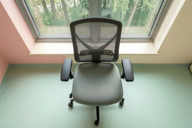 Een lege ergonomische bureaustoel staat naar een groot raam met groene bomen gericht in een kamer met pastelroze muren en een mintgroene vloer.