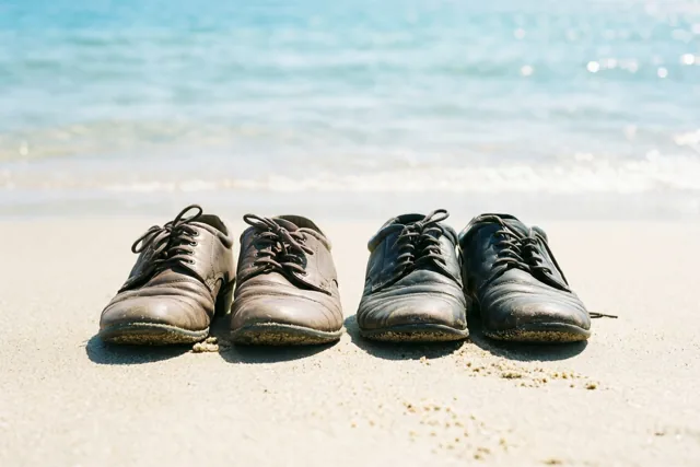 Twee paar versleten leren schoenen staan naast elkaar op het zandstrand aan de waterlijn, met blauw zeewater op de achtergrond in fel zonlicht.