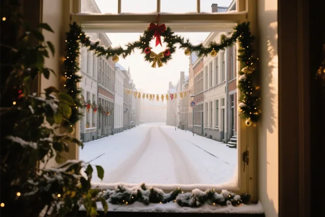 Door een raam versierd met kerstslingers en sneeuw is een lege, besneeuwde straat in Leuven te zien in zacht warm licht.