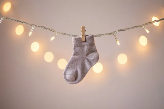 Een lichtgekleurd babysokje zit met een knijper vast aan een lichtsnoer, voor een wazige achtergrond met warme lichtcirkels.