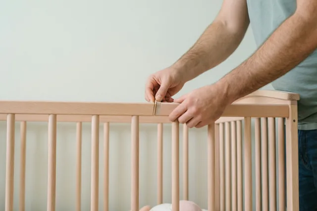De handen van een man zetten een babybedje van licht hout in elkaar tegen een effen muur, bij zacht daglicht.