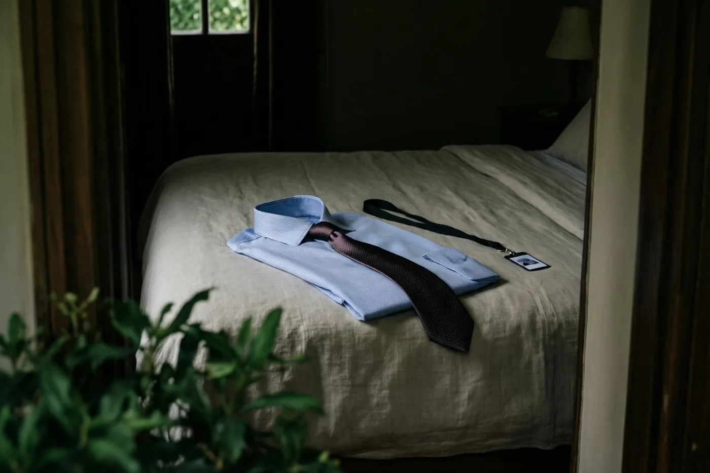 Door een houten deuropening zijn een opgevouwen lichtblauw hemd, een donkere das met patroon en een zwart keycord met een identificatiekaart te zien op een beige linnen bed in zacht daglicht.