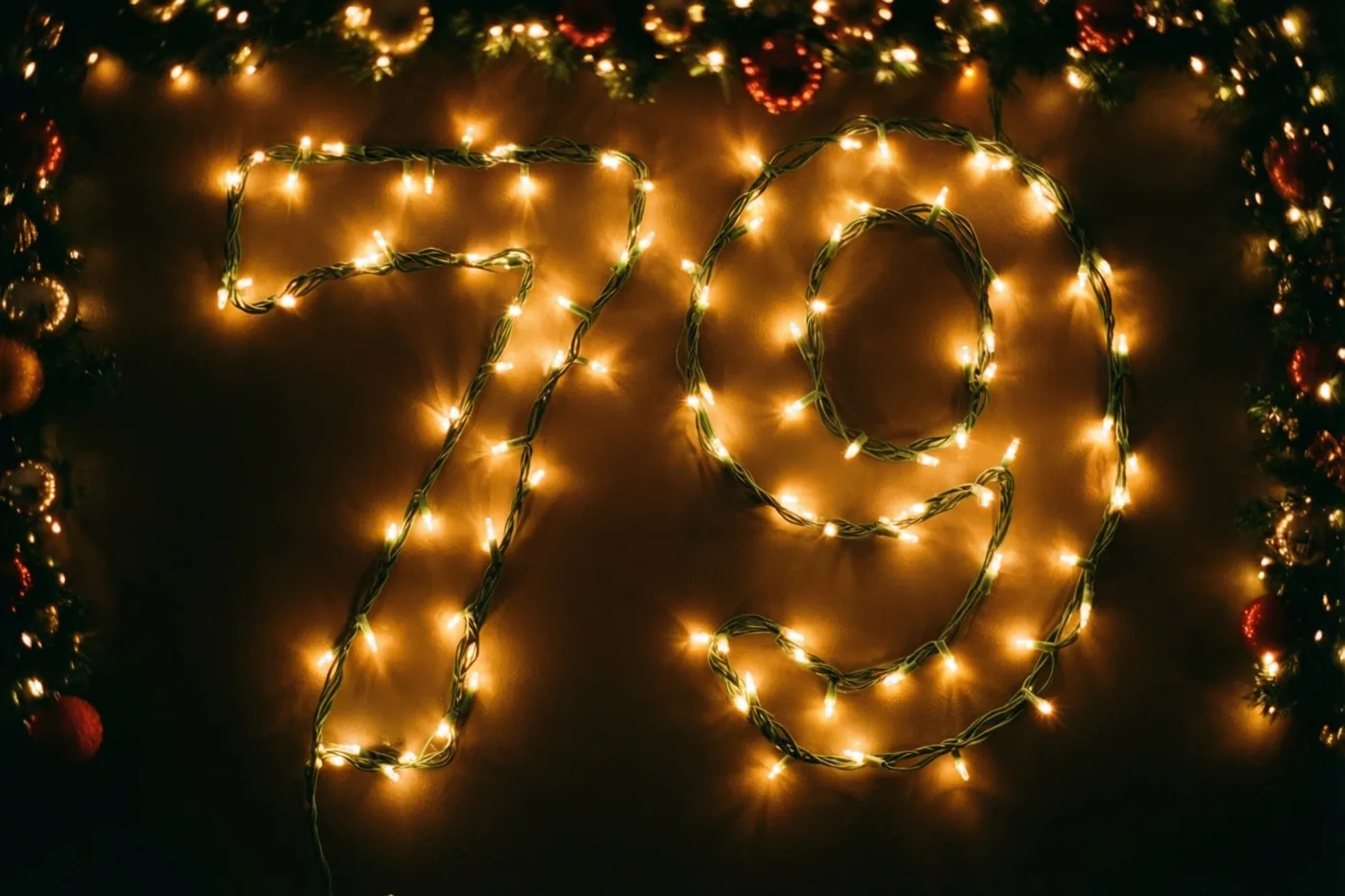 Het getal 79 van warme lichtslingers hangt tegen een donkere muur, omlijst door een kerstslinger en onscherpe lichtjes in gouden en rode tinten.