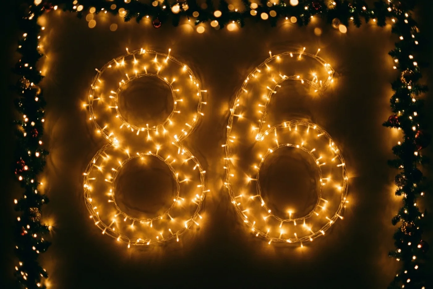Warme lichtjes vormen 86 tegen een donkere muur, omlijst door een decoratieve slinger met onscherpe lichtjes op de achtergrond.