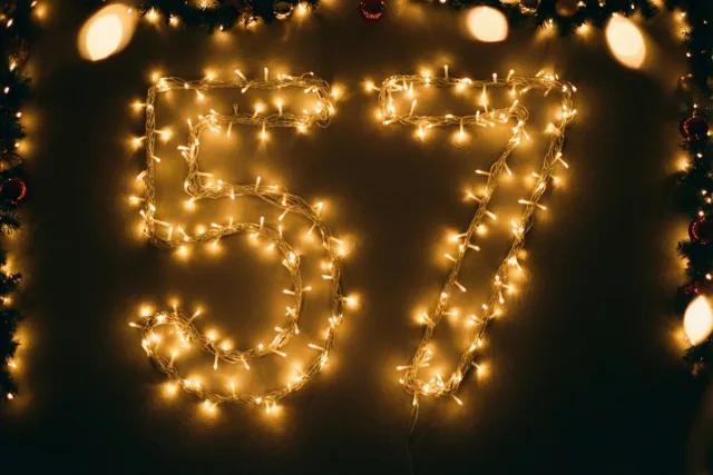 Warm gloeiende lichtjes vormen 57 op een donkere muur, omringd door onscherpe feestlichtjes en een dennenslinger.