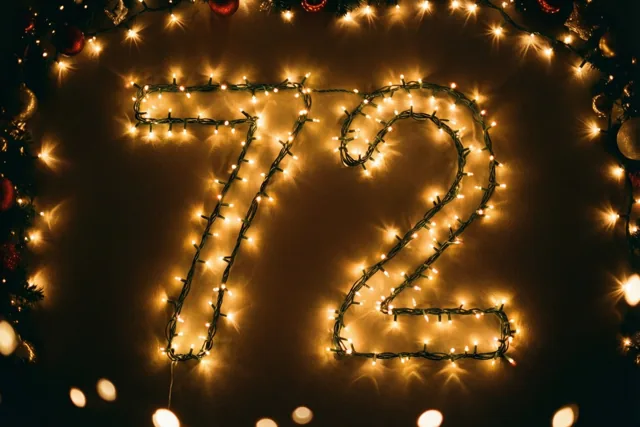 Warme lichtjes vormen 72 op een donkere muur, omringd door onscherpe lichtpunten en feestversiering.