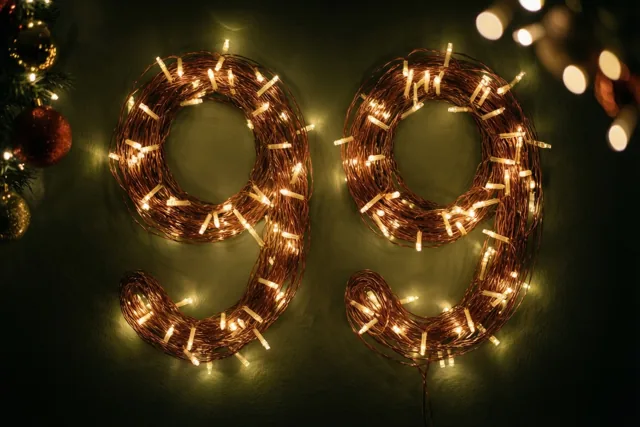 Warme lichtslingers vormen 99 tegen een donkere muur, met onscherpe lichtjes en kerstversieringen in de hoeken.