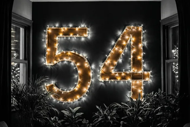 Warme goudkleurige lichtjes vormen 54 op een donkere muur, omlijst door ramen en potplanten.