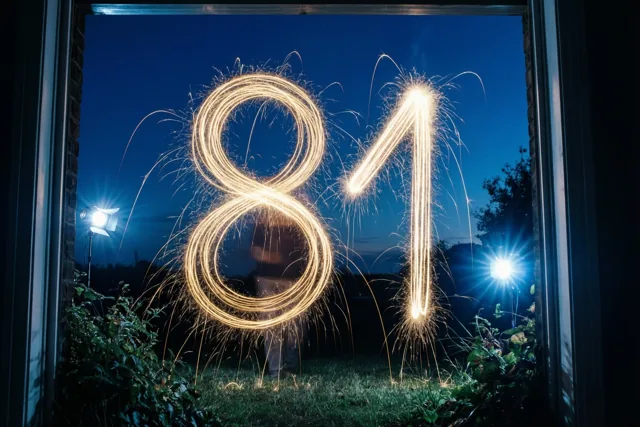 Gouden lichtsporen van sterretjes vormen 81 tegen een donkerblauwe nachthemel, gezien door een raamkozijn met felle schijnwerpers aan weerszijden.