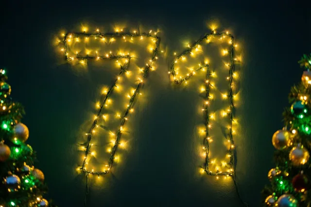 Warme lichtjes vormen 71 op een donkere muur, met aan weerszijden onscherpe kerstbomen met versiering.