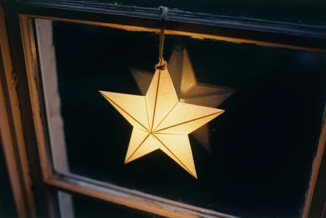 Een lichtgevende papieren ster hangt aan een touwtje in een donker raam met een houten kader, met een lichte rand rond de foto.