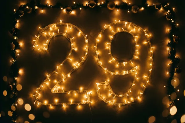 Warme lichtjes vormen het getal 29 tegen een donkere muur, met een slinger en onscherpe lichtvlekken eromheen.