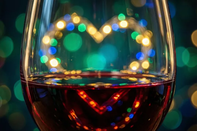 Een close-up toont rode wijn in een glas met een hartvormig patroon van onscherpe lichtslingers erdoorheen en kleurrijke lichtvlekken op een donkere achtergrond.