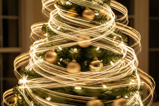 Een kerstboom met groene takken en gouden versiering staat binnen tegen een schemerige achtergrond, met kringelende gouden lichtstrepen van een langzaam draaiende lichtbron.