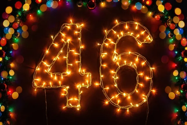 Warme lichtslingers vormen het getal 46 tegen een donkere muur, omringd door onscherpe lichtjes in goud, rood en groen.