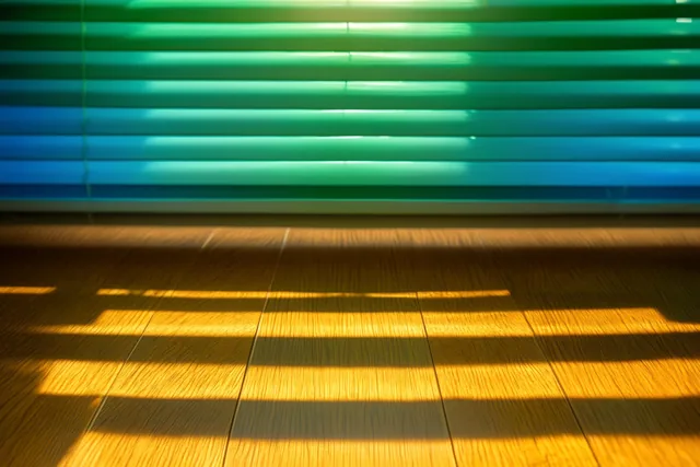 Zonlicht valt door horizontale jaloezieën met groene en blauwe tinten en tekent warmgele strepen op een houten vloer.