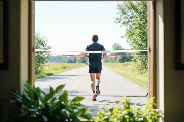 Door een raam met groen op de voorgrond is een loper van achteren te zien die op een open weg door een finishlint loopt, in helder zonlicht.