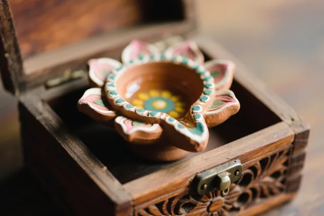 Een handbeschilderd lotusvormig olielampje van klei ligt in een open houten doos met snijwerk, in warme aardetinten met roze, blauwgroene en gele accenten.
