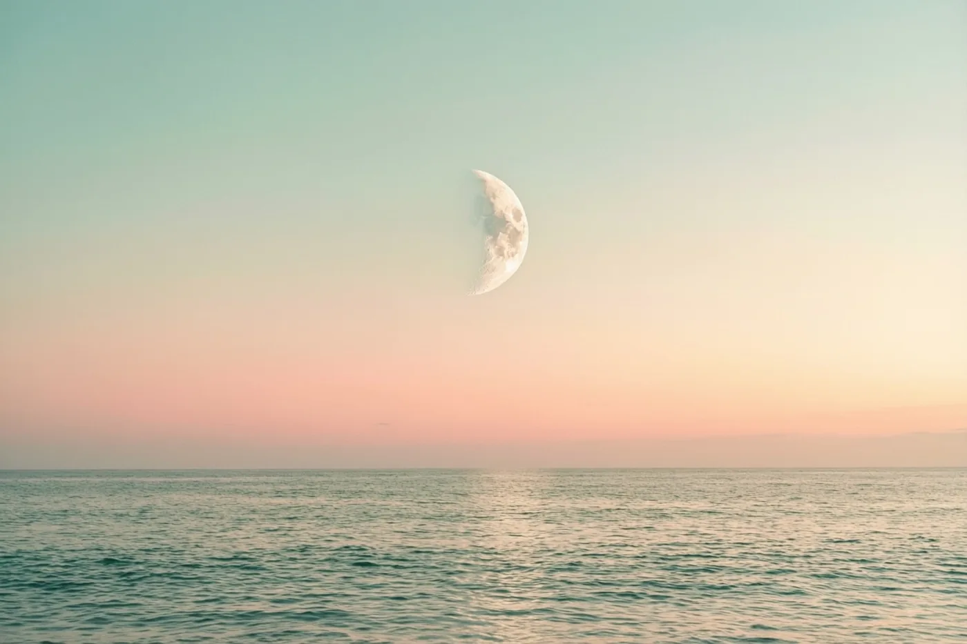Een maansikkel staat boven een kalme zee bij zonsopgang, onder een hemel die van roze naar blauwgroen overgaat en zacht weerspiegelt in het water.