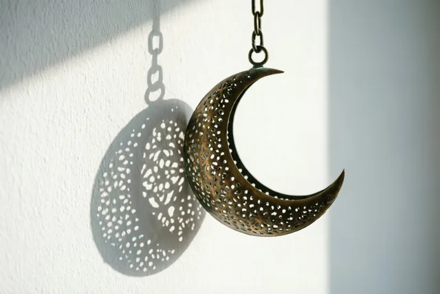 Een messing maansikkellantaarn met fijne opengewerkte patronen hangt aan een ketting en werpt in natuurlijk zonlicht een gedetailleerde schaduw op een witte muur.