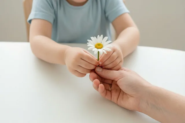 Kinderhanden reiken boven een witte tafel een wit madeliefje met een geel hart naar de hand van een volwassene, tegen een neutrale achtergrond in zacht licht.