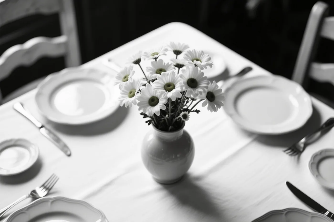 Een zwart-witfoto toont een vaas met madeliefjes midden op een gedekte ontbijttafel met borden en bestek, met sterke contrasten en zachte schaduwen.