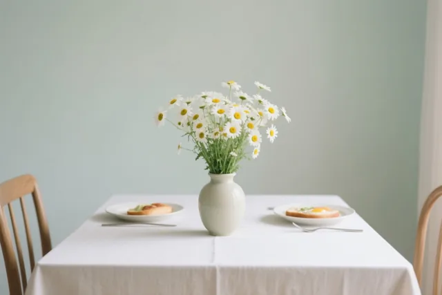 Een witte vaas met madeliefjes staat op een witgedekte ontbijttafel tussen twee borden met eten en vorken, in een eetkamer met lichtgroene muren en houten stoelen.