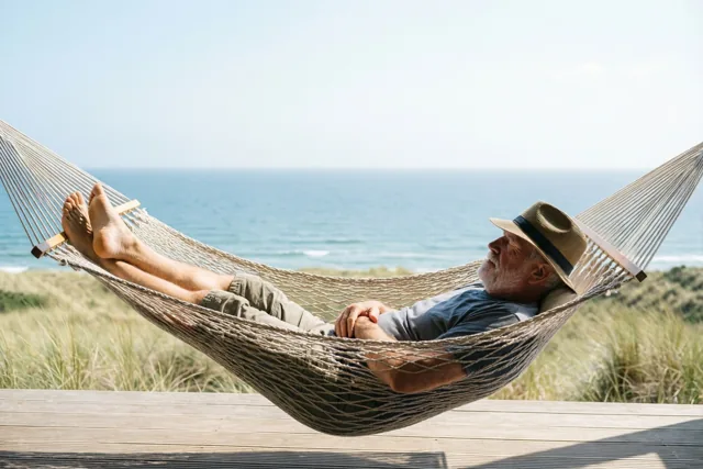 Een oudere man met een witte baard en strohoed dommelt in een beige touwhangmat boven een houten terras in een zonnig kustduin, met een blauwe zee onder een heldere hemel.