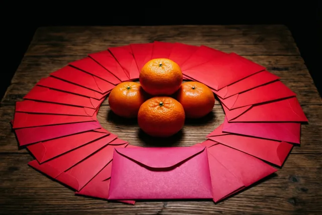 Vier mandarijnen liggen in een kring op een houten tafel, omringd door uitgewaaierde rode enveloppen in warm licht tegen een donkere achtergrond.