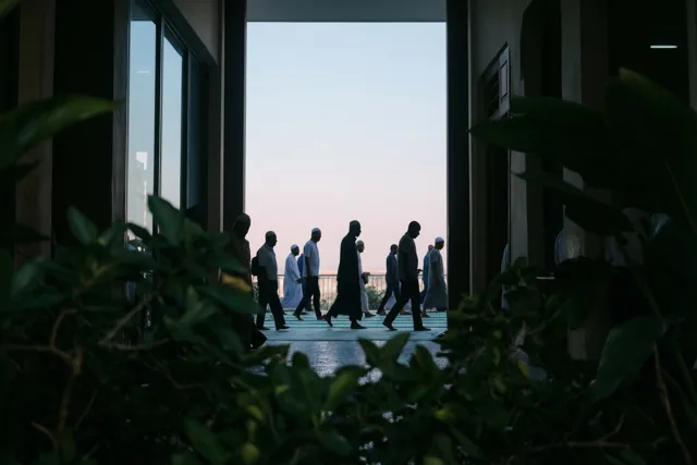 Silhouetten van mannen lopen achter elkaar naar een lichte opening onder een bleke hemel, met donker gebladerte op de voorgrond.