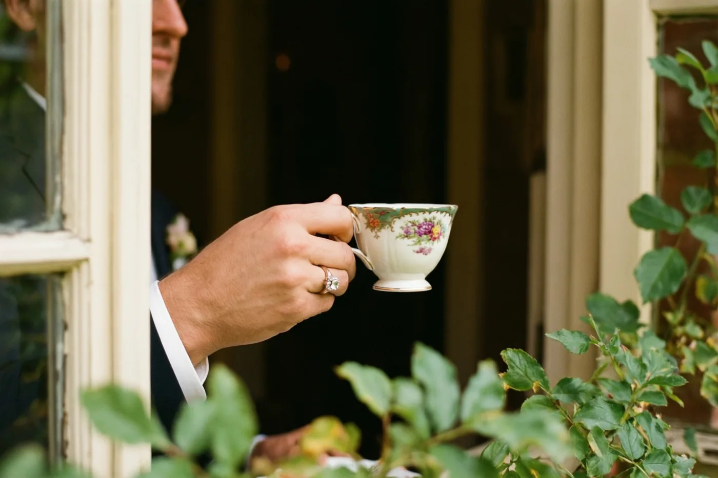 Een mannenhand met een verlovingsring houdt een vintage theekopje met bloemen vast, met raamstijlen rondom en groen op de voorgrond.